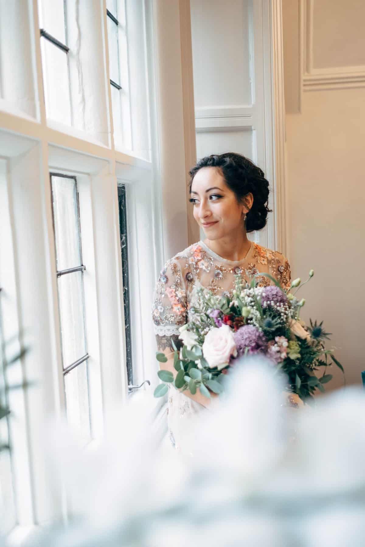 bride looks out of window holding her bouquet at her lewes diy village hall wedding
