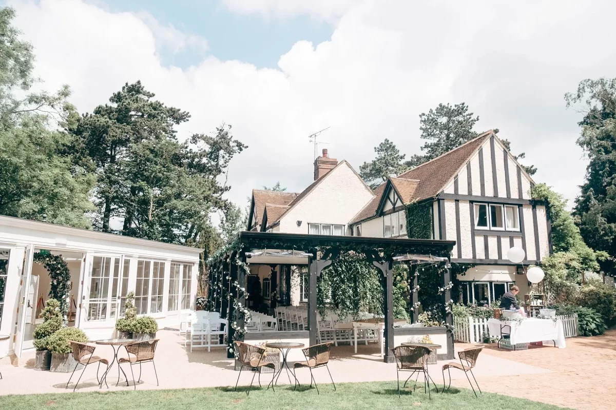 A charming country house with ivy-covered walls and Tudor-style architecture, perfect for hayne house wedding photography, featuring outdoor seating on a patio beneath a pergola, surrounded by lush greenery and tall trees.