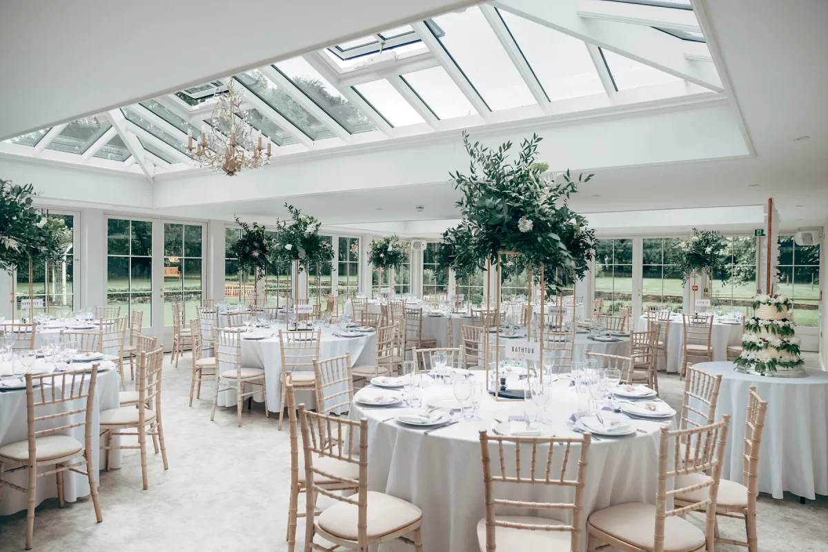 Elegant wedding reception room at Hayne House with round tables draped in white cloths, wooden chairs, towering green floral centerpieces, and natural light streaming through a glass ceiling—perfect for stunning Hayne House wedding photography. A tiered cake sits to the right.