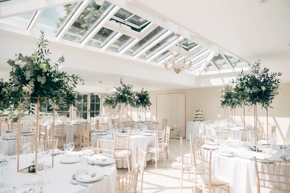 A bright, elegant event space with large skylights, white walls, round tables set for dining, gold chairs, and tall floral centerpieces—perfect for capturing stunning hayne house wedding photography in an airy and sophisticated atmosphere.