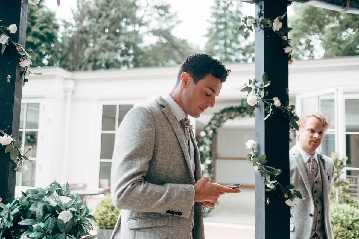 A man in a light grey suit checks his phone under a floral-decorated archway, while another stands nearby. Captured at an outdoor event with greenery and white flowers, this scene reflects the elegance of Hayne House wedding photography.