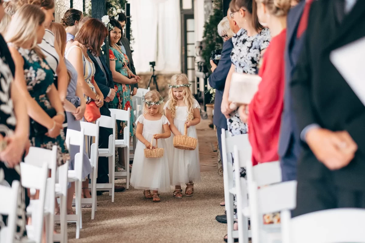 Two young flower girls in white dresses and flower crowns walk down the aisle holding baskets, surrounded by smiling wedding guests. This charming moment is beautifully captured in our Hayne House wedding photography.
