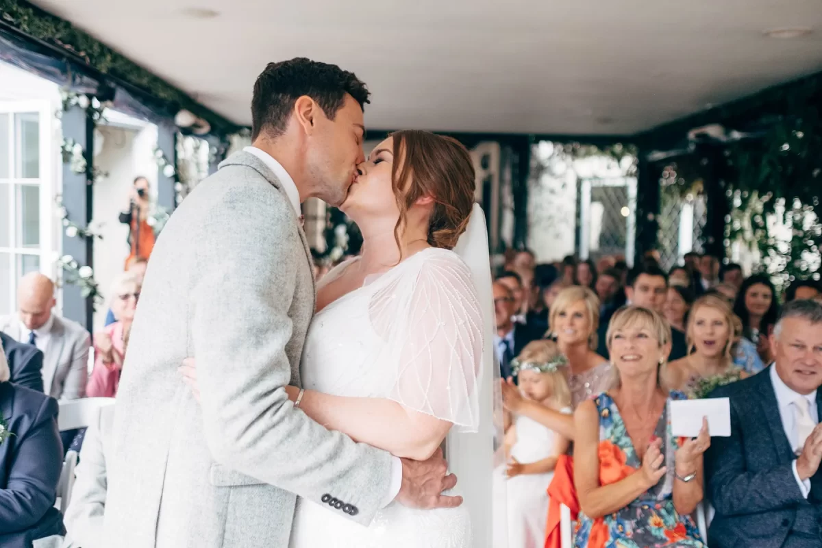 A bride and groom kiss at their wedding ceremony, surrounded by smiling guests who applaud and capture the moment. The setting is bright, decorated with greenery—a perfect scene for Hayne House wedding photography.