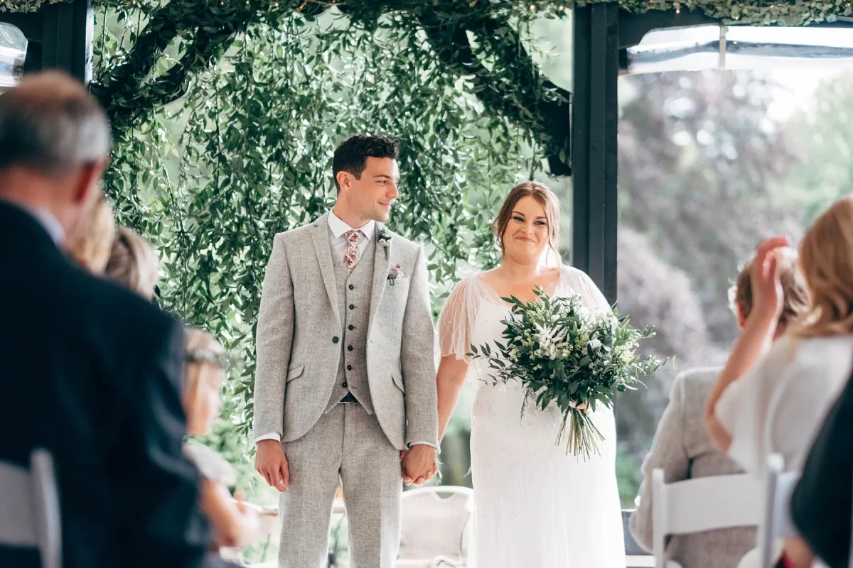 A bride and groom stand holding hands at their wedding ceremony, smiling at each other. With lush foliage and greenery in her bouquet, the moment is beautifully captured in this Hayne House wedding photography scene as guests look on.
