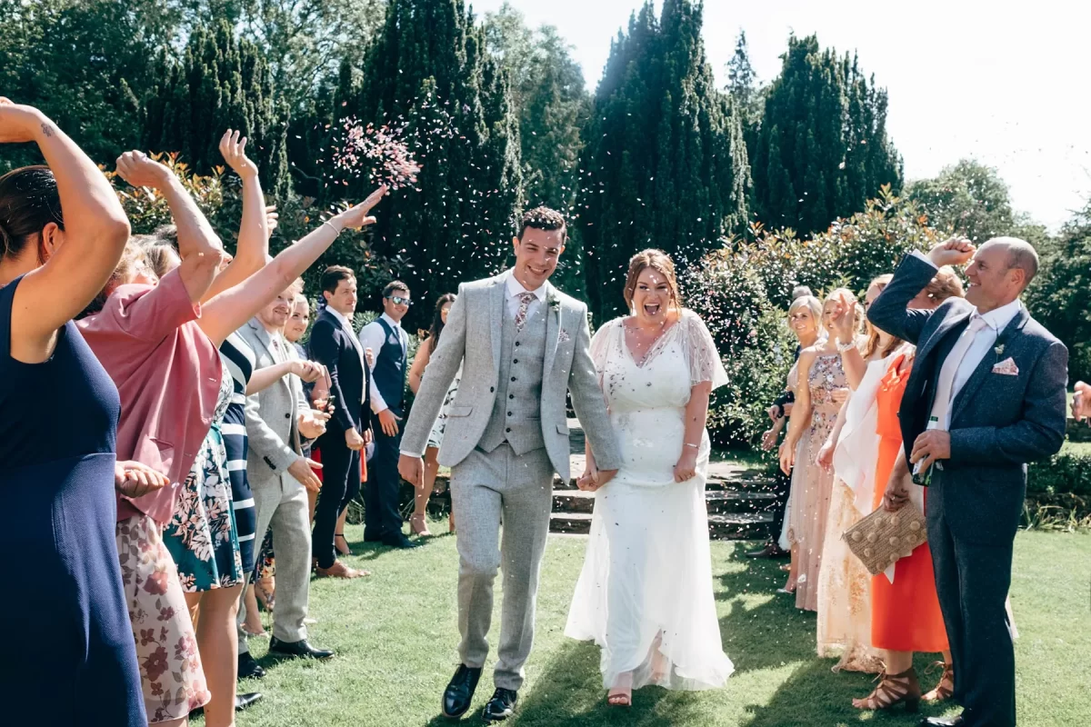 A bride and groom walk outdoors, smiling and holding hands, as guests on both sides throw confetti to celebrate their wedding on a sunny day—captured beautifully with Hayne House wedding photography.
