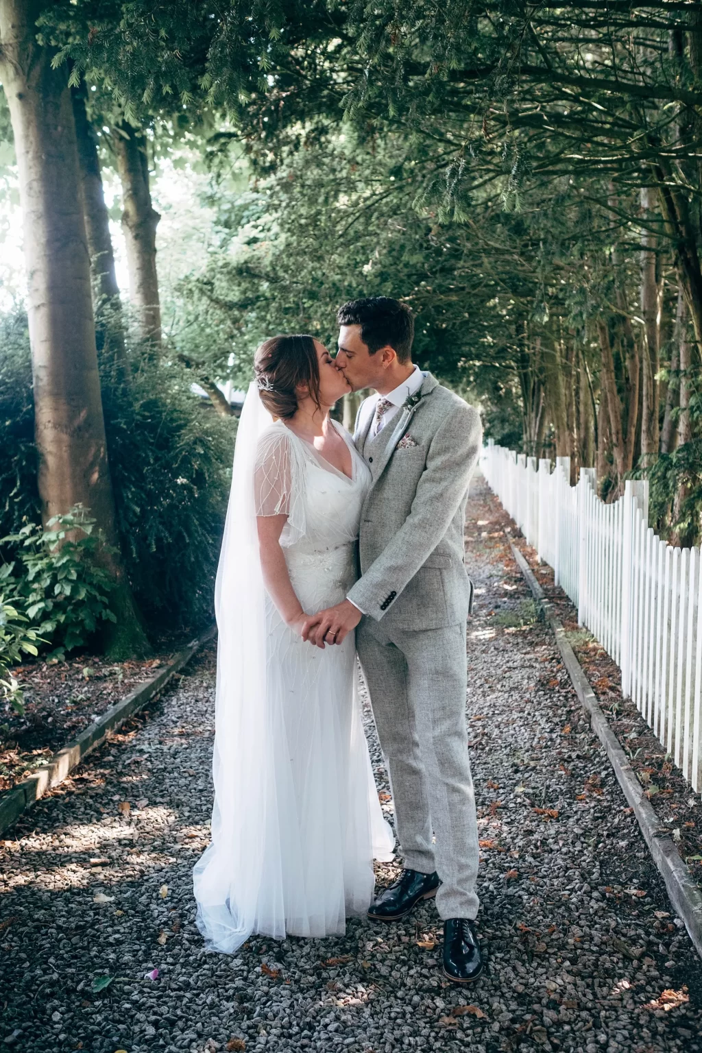 A bride and groom, dressed in a white gown and light gray suit, share a kiss on a tree-lined gravel path with a white fence—captured beautifully in this Hayne House wedding photography moment.