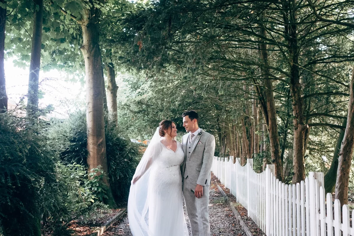 A bride and groom stand close together on a wooded path next to a white picket fence, surrounded by tall trees. Capturing their loving gaze, this scene beautifully showcases romantic Hayne House wedding photography.