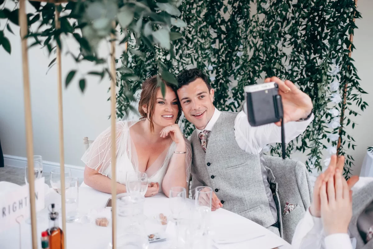 A smiling couple dressed in wedding attire sits at a decorated table, posing for a selfie together. Greenery fills the background, and empty glasses are on the table—classic moments captured with Hayne House wedding photography.