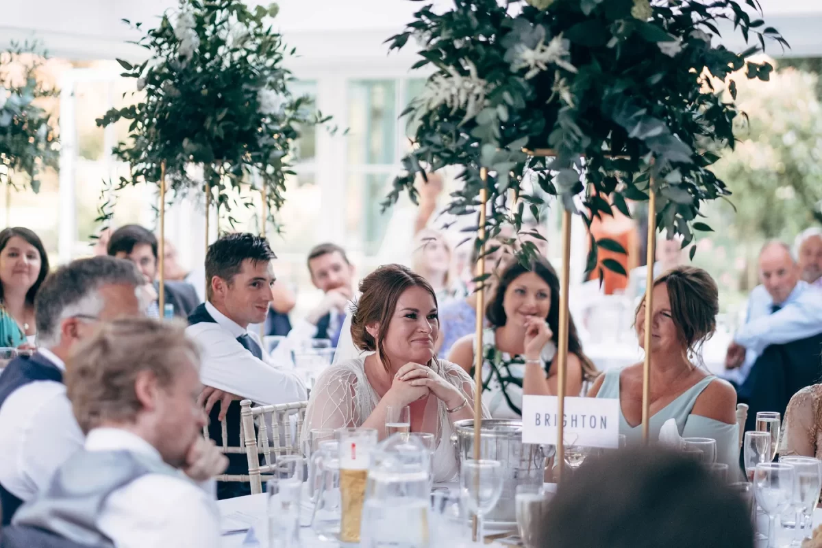 A group of people dressed formally sit at round tables decorated with tall green floral centerpieces, attentively listening and smiling during what appears to be a wedding reception—an elegant moment captured in Hayne House wedding photography.