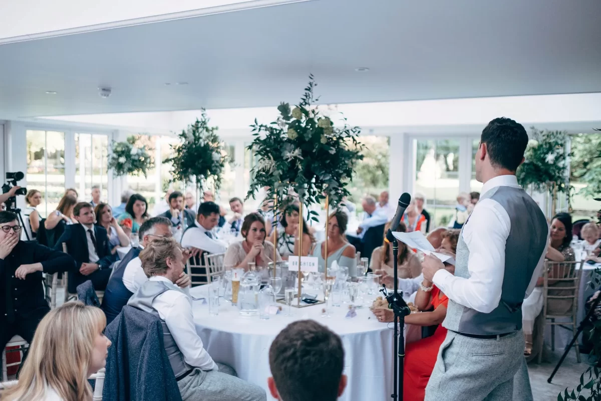 A man in a vest gives a speech at a wedding reception, standing by a microphone and holding notes, while guests seated at round tables listen attentively in a bright, decorated venue—perfect for Hayne House wedding photography.