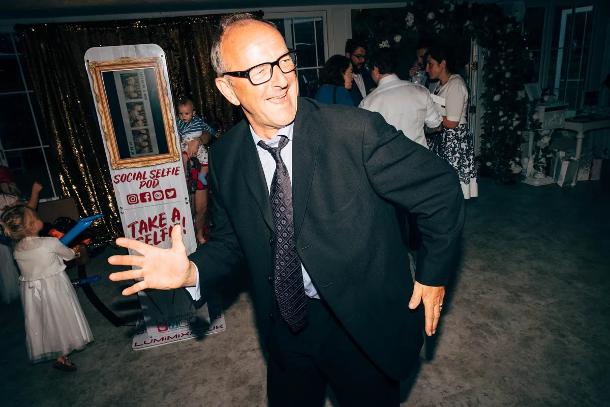 A man in a suit and glasses dances with a smile at a lively indoor event, with people and children mingling near a selfie booth—capturing the joyful spirit of Hayne House wedding photography.