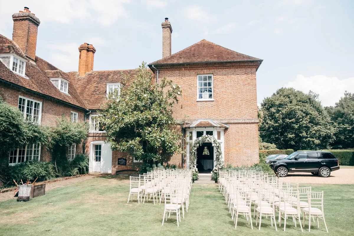 Rows of white chairs are set up on a lawn facing the entrance of a large, historic brick house with greenery and a black SUV parked nearby, ready for an outdoor event or wedding ceremony.