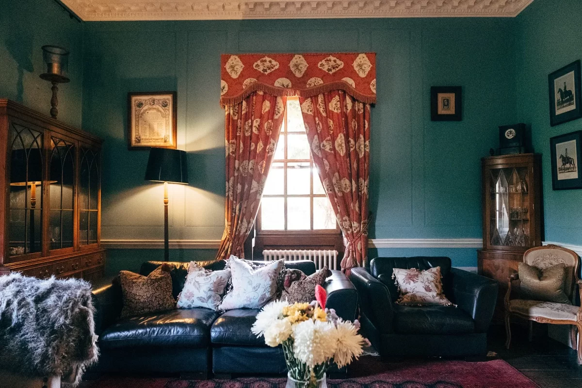 A cozy living room with teal walls, a black leather sofa with assorted pillows, a window with red and gold patterned curtains, vintage cabinets, framed art, and a vase of white flowers on a table in the foreground.
