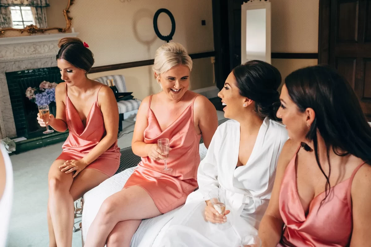 Four women sit on a bed, laughing and talking. Three wear matching pink satin dresses, and one wears a white robe. They each hold a glass of champagne in a warmly lit, elegant room.