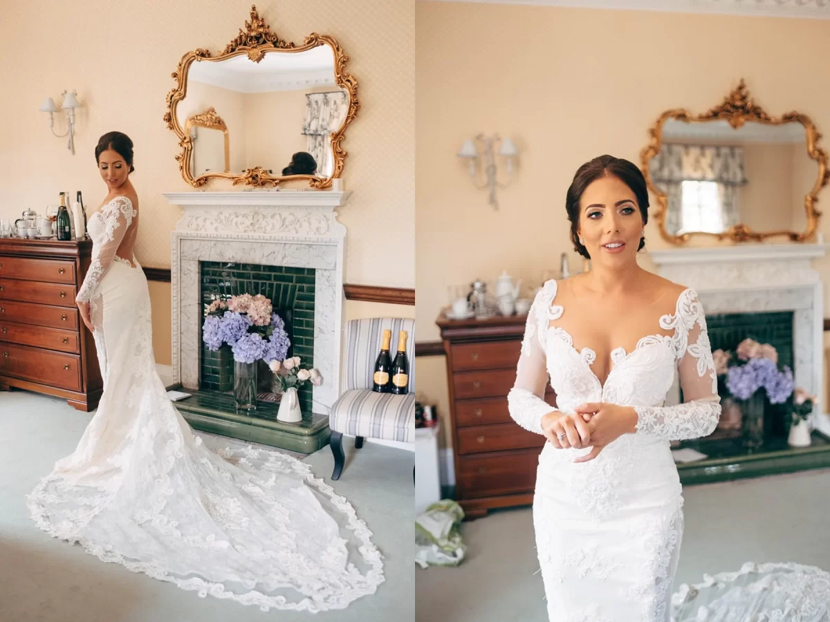 A bride in a white lace gown with a long train stands and poses in an elegant room with a fireplace, mirror, flowers, and vintage decor.