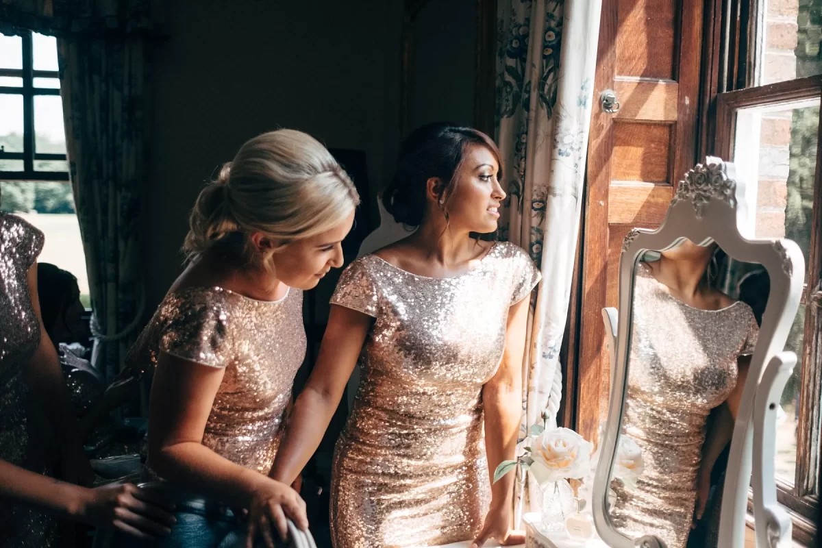Two women in sparkly, matching dresses stand by a window, looking outside. One smiles while holding the other’s hand. Sunlight streams in, illuminating them and their reflections in a mirror on the table nearby.