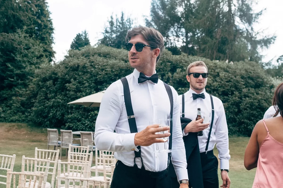 Two men wearing white shirts, black suspenders, bow ties, and sunglasses stand outdoors on grass. One holds a glass, the other a bottle. Empty chairs and greenery are in the background. A woman in a pink dress is partially visible.