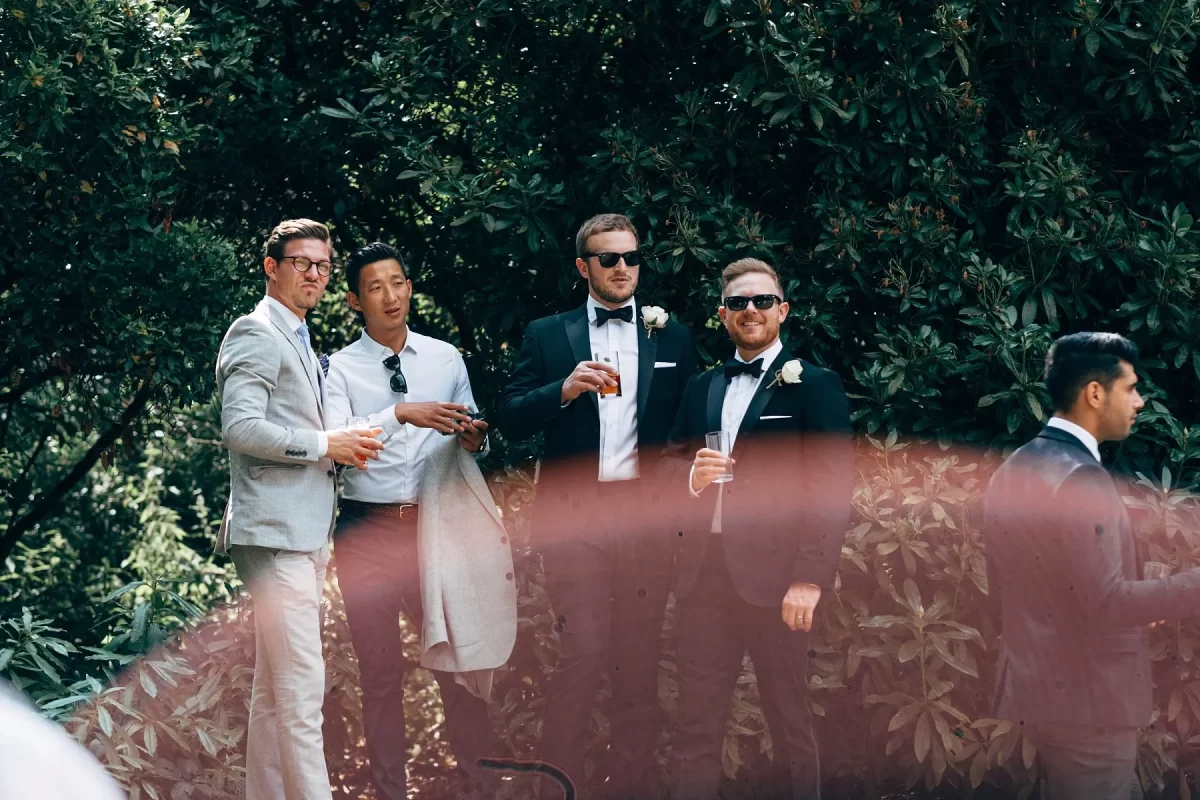 Four men dressed in suits stand outdoors, holding drinks and posing for a photo in front of leafy green bushes. Another man stands to the right, slightly apart. A blurry pinkish shape appears across the foreground.