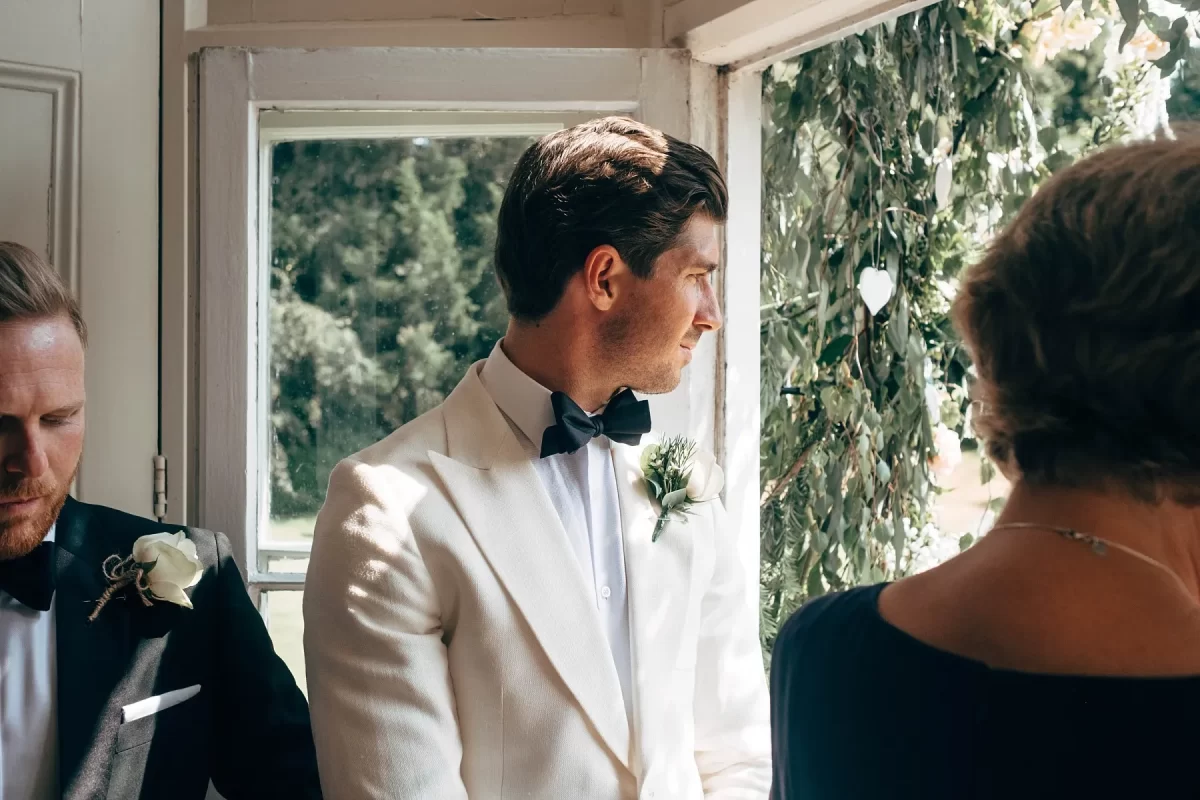 A man in a white tuxedo and black bow tie stands indoors, looking thoughtfully out a sunlit window decorated with greenery. Another man and a woman are partially visible beside him.