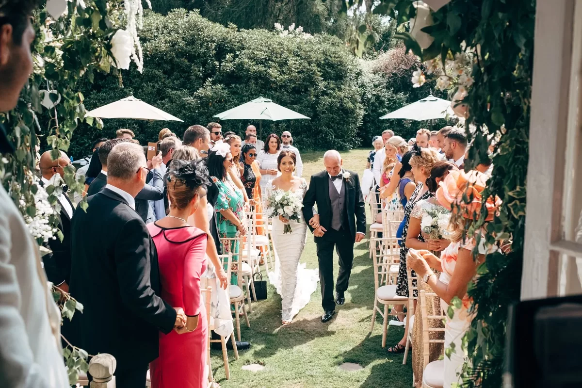 A bride in a white dress walks down an outdoor aisle with an older man, surrounded by seated guests in formal attire. The setting is lush with greenery and decorated with flowers and parasols.