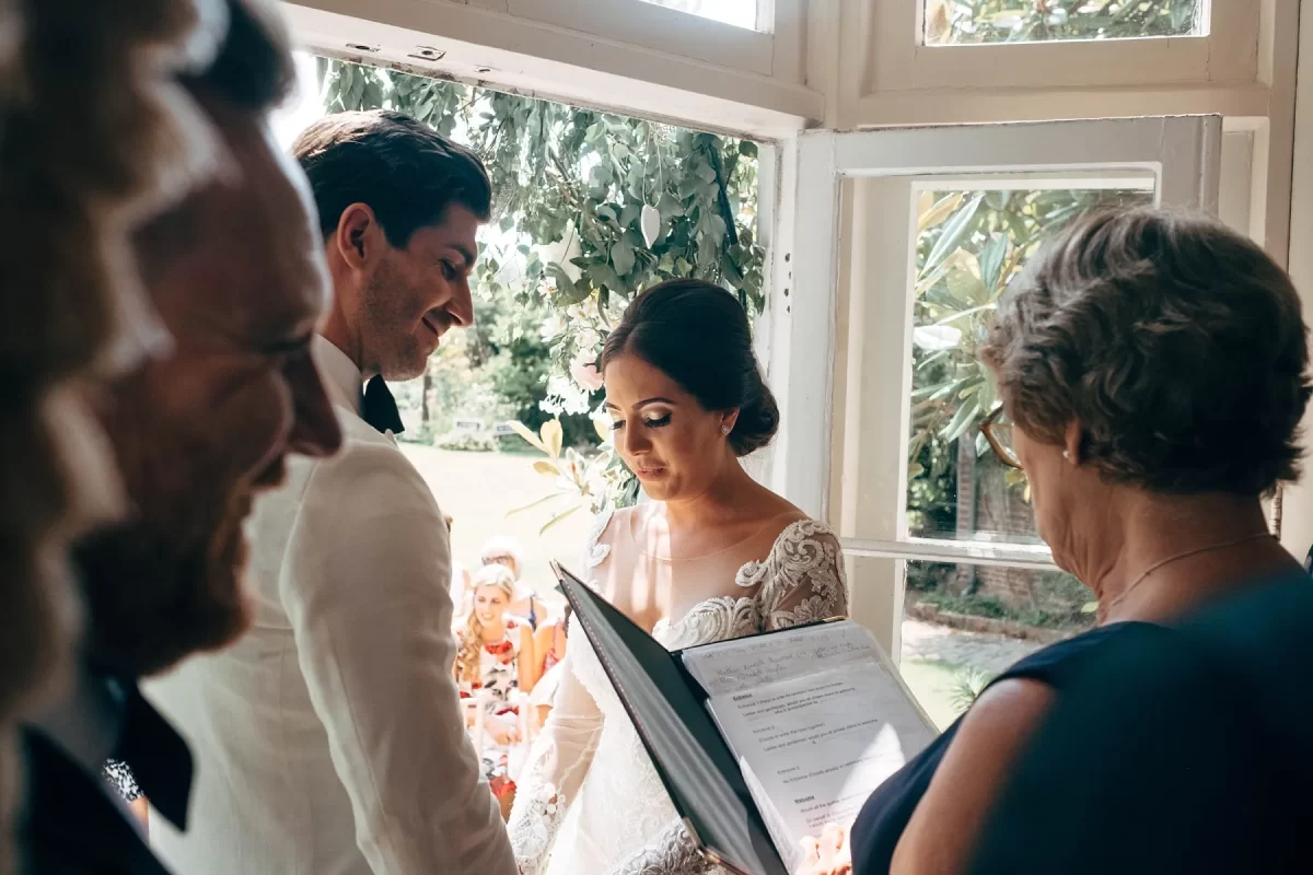 A bride and groom stand facing each other, smiling, during a wedding ceremony by a sunlit window, surrounded by officiant and guests, with greenery visible outside.
