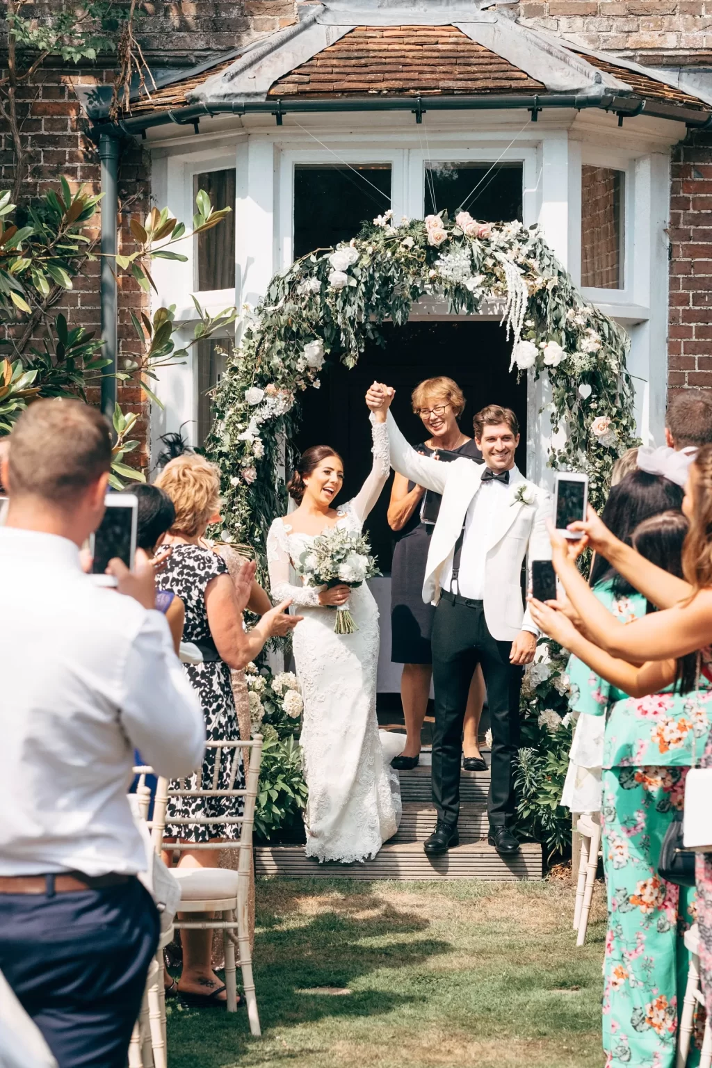 A newlywed couple, smiling and holding hands, walks under a floral arch as guests take photos and cheer. The bride wears a white dress and holds a bouquet; the groom wears a white jacket and black trousers at Sprivers Mansion