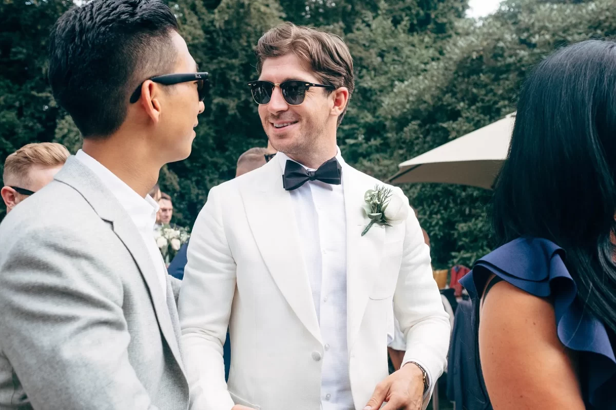 Two men in sunglasses chat and smile at an outdoor event. One wears a white suit with a black bow tie and boutonniere, suggesting a formal occasion like a wedding. Greenery and a large umbrella are visible in the background.