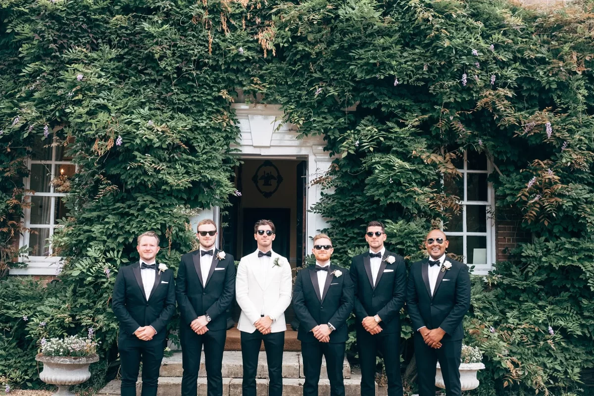 Six men in tuxedos, with one in a white jacket and the others in black, stand in a row outside a building covered with green vines, posing for a formal photo on steps in front of a doorway.