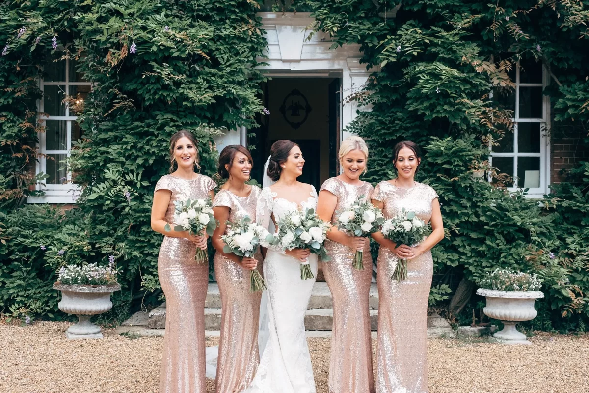 Five women in elegant dresses, one in white and four in matching sparkling gowns, stand together holding bouquets in front of a green, vine-covered building with white trim. They are smiling and posing for a photo.