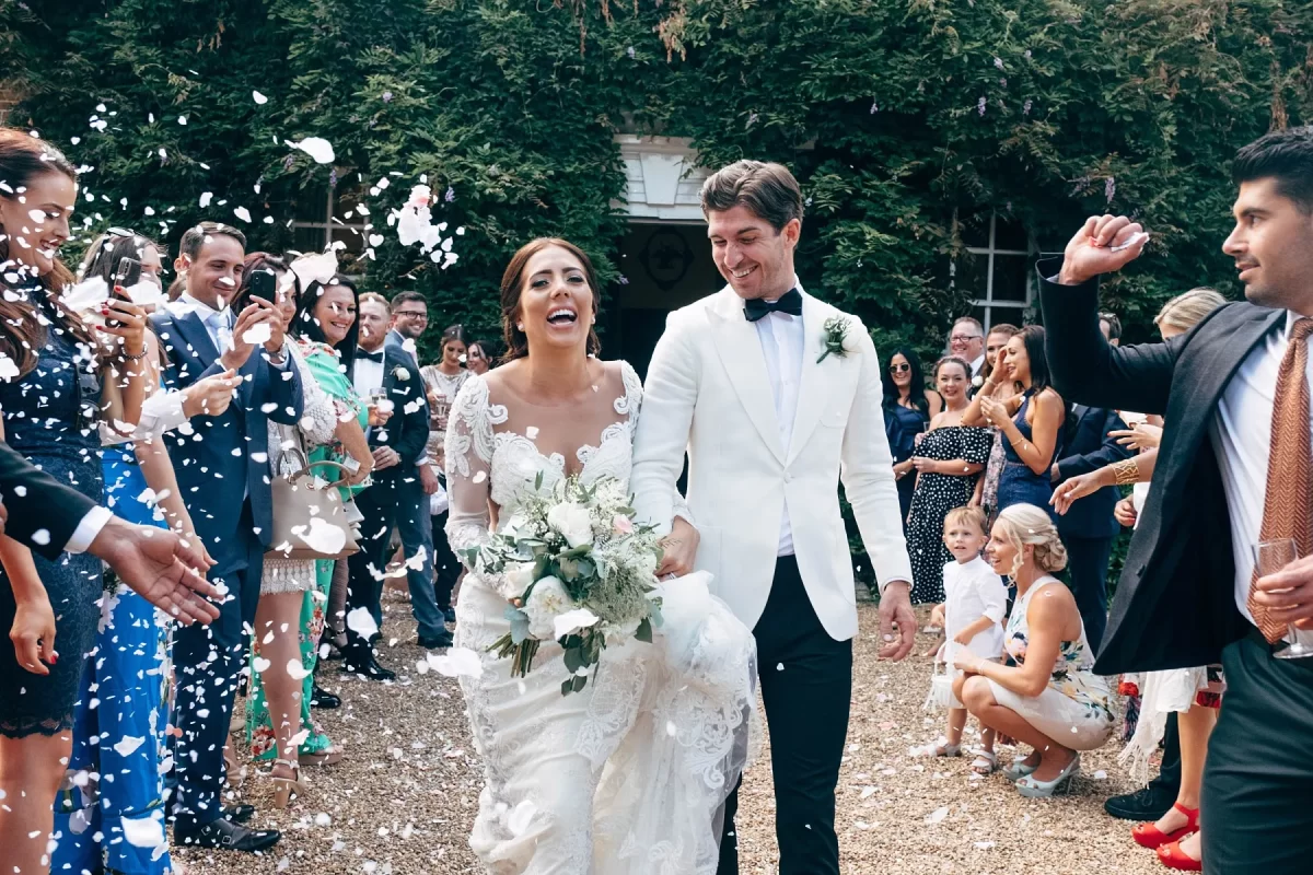 A bride and groom walk hand-in-hand, smiling joyfully, as guests on both sides throw white confetti. The couple is dressed elegantly, and the outdoor setting is lush with greenery and celebratory guests.