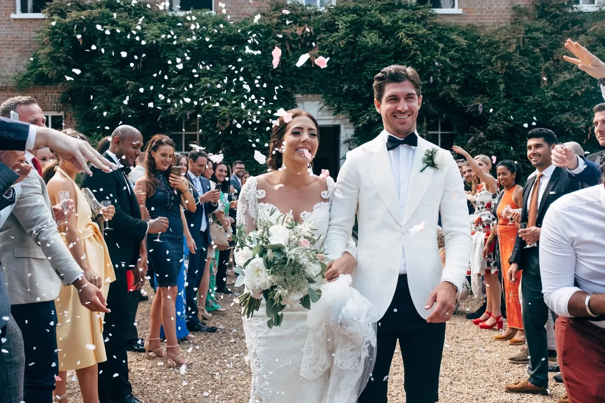 A bride and groom in formal attire walk outdoors, smiling, as guests throw confetti and cheer. The bride holds a bouquet of flowers and the groom wears a white tuxedo jacket. People celebrate around them in colorful outfits.