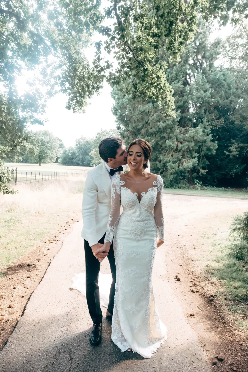 A groom in a white jacket kisses the smiling bride on the cheek as they hold hands and walk down a sunlit tree-lined path, surrounded by greenery on their wedding day.