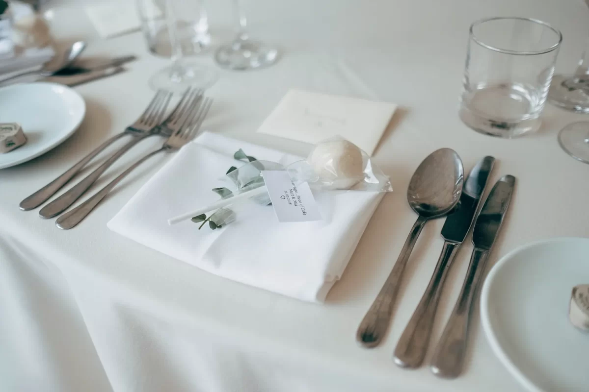 A formal table setting with utensils, a white napkin, a small sprig of greenery, a wrapped round favor, a glass, and a folded card on a white tablecloth.