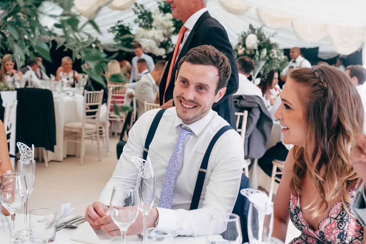 A smiling man in a shirt and suspenders sits at a decorated table beside a woman, both surrounded by guests at a formal event, likely a wedding reception, in a bright, elegant venue.