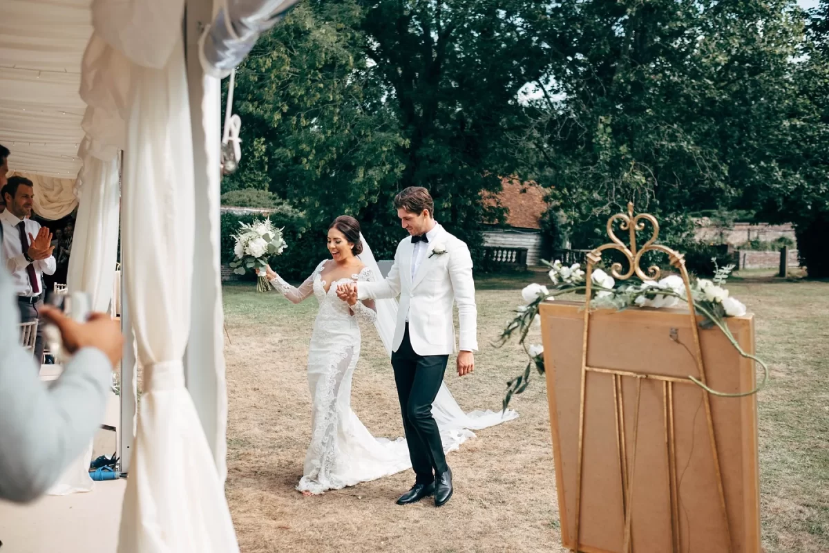 A bride and groom, holding hands and smiling, walk into an outdoor wedding reception tent. The bride wears a white gown and holds a bouquet; the groom wears a white jacket and black pants. Guests are applauding.