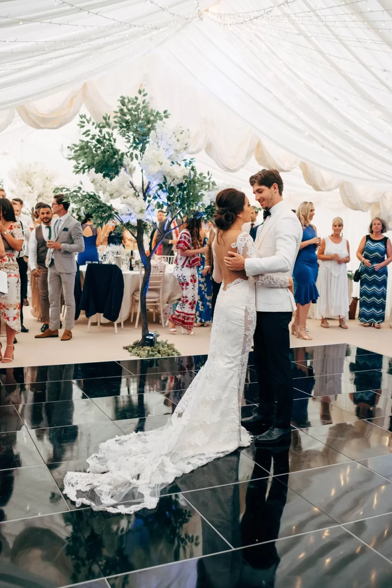 A bride and groom share their first dance on a black-tiled floor under a white tent, surrounded by wedding guests and white floral decorations.