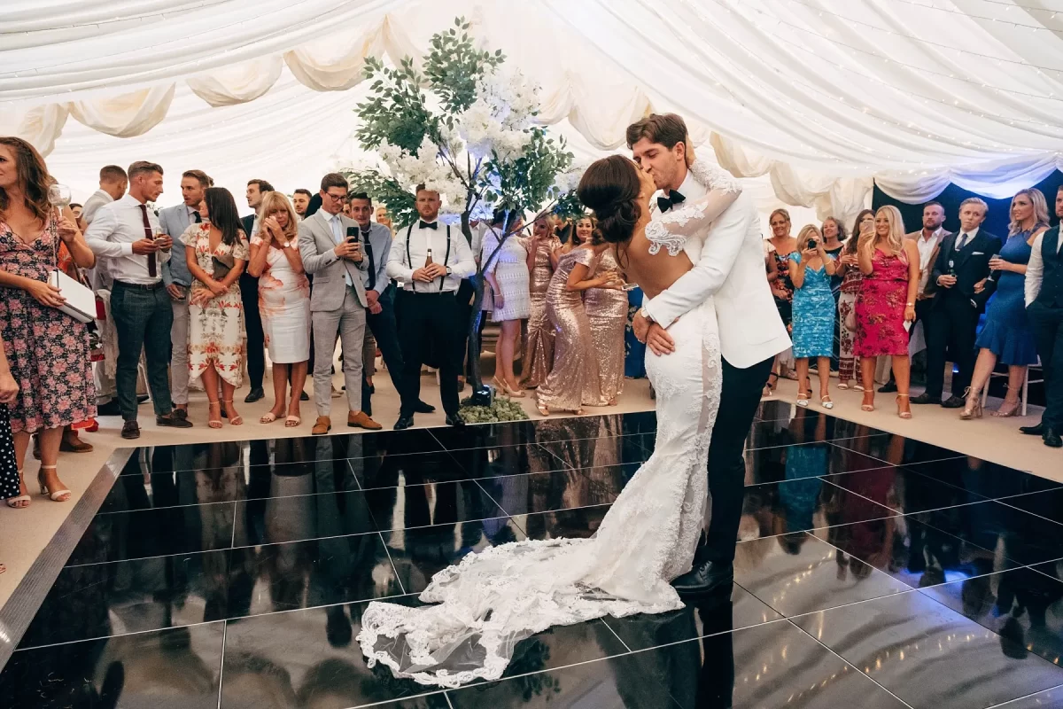 A bride and groom share their first dance on a black dance floor under a white tent, surrounded by elegantly dressed guests watching and smiling.
