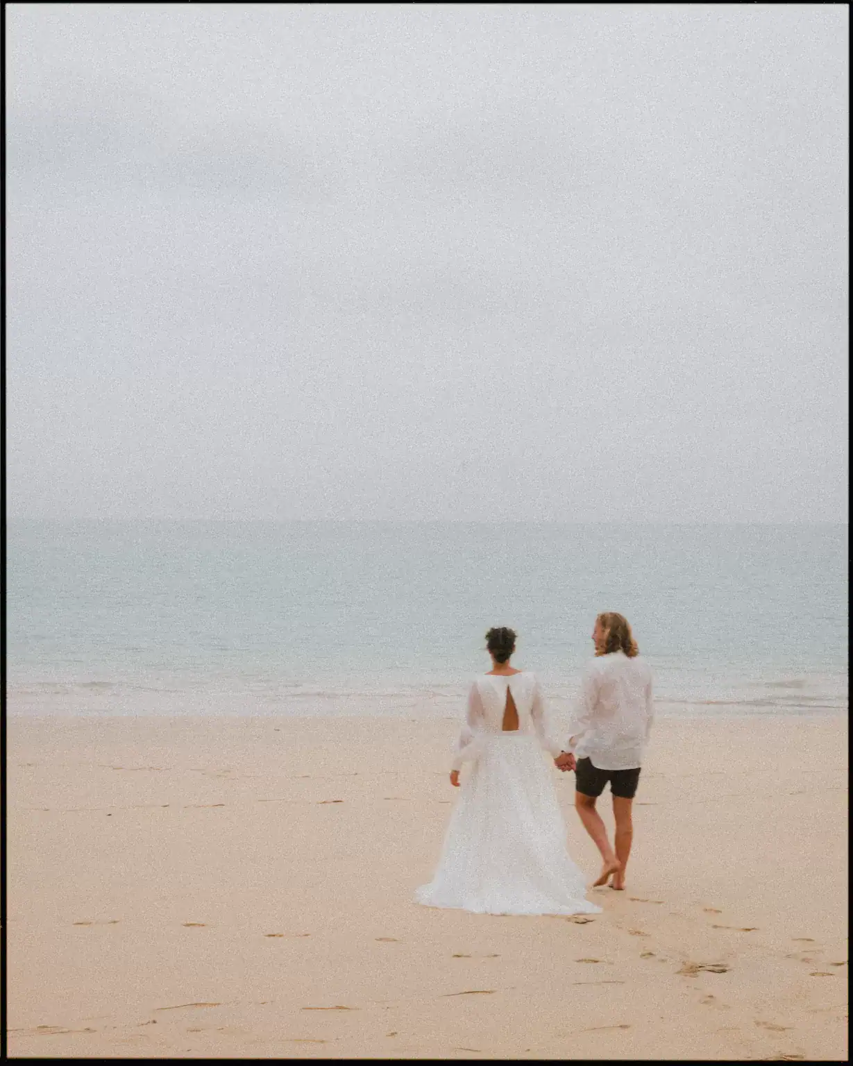 A couple stands hand in hand on a sandy beach near St Ives Harbour Hotel, facing the calm sea. The person on the left wears a long white dress; the person on the right wears a white shirt and black shorts. The sky is overcast.