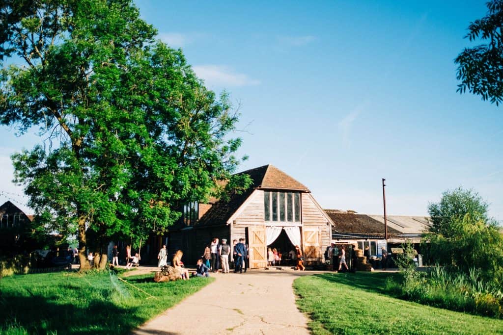 A rustic barn wedding with open wooden doors hosts a gathering of people outside on a sunny day, surrounded by green grass, trees, and clear blue sky.