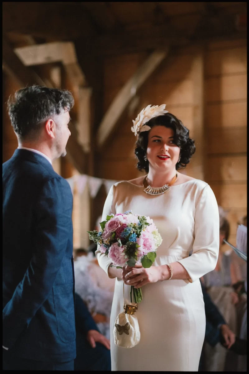 A bride in a white dress holding pink and purple flowers faces a groom in a suit during a wedding ceremony at the rustic, wood-paneled Yoghurt Rooms wedding festival venue.