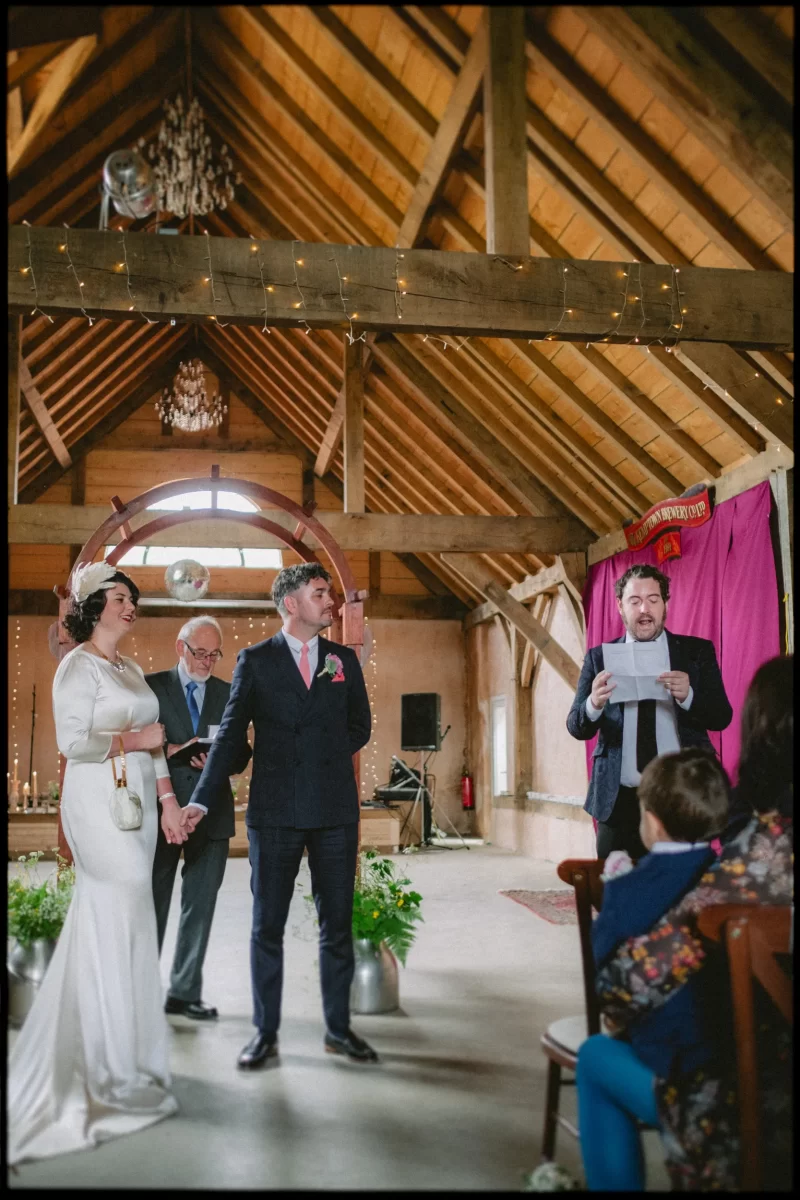 A wedding ceremony at the Yoghurt Rooms, with a rustic barn festival vibe. The bride and groom hold hands and smile as a man reads from a paper. Guests are seated beneath fairy lights strung across the wooden beams above.