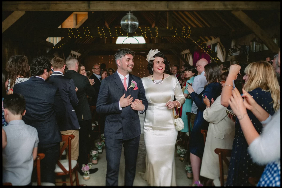 A newlywed couple walks down the aisle smiling at a Yoghurt Rooms wedding festival, surrounded by cheering guests in a rustic, warmly lit venue decorated with string lights and flowers. The bride wears a white dress and fascinator; the groom a dark suit.