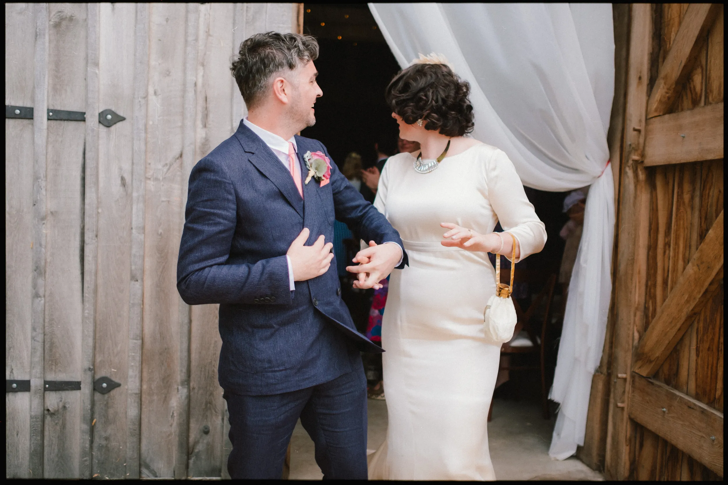 A man in a navy suit and a woman in a long white dress holding a purse smile and hold hands as they walk out of a wooden barn door draped with white fabric at their yoghurt rooms wedding festival.