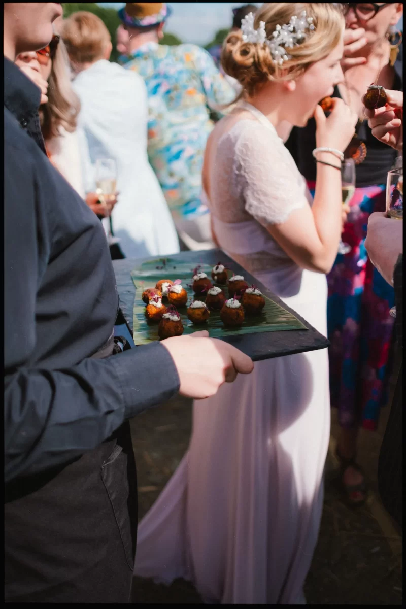 A server in black holds a tray of bite-sized appetizers at an outdoor wedding festival. In the background, a woman in a white dress and floral headpiece is eating, surrounded by guests in colorful outfits near quaint yoghurt rooms.