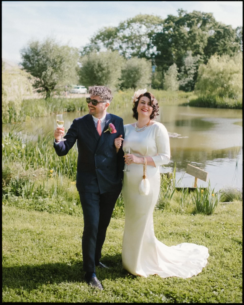 A smiling bride and groom walk on grass beside a pond at a Yoghurt Rooms wedding festival, holding champagne glasses. The groom wears a dark suit and sunglasses, while the bride shines in a long white dress and pearl necklace amid lush greenery.