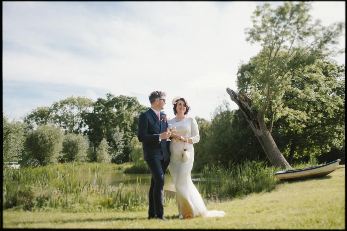A bride and groom smile outdoors on the grassy lawn at their Yoghurt Rooms wedding festival, holding glasses near a pond. Trees and a small boat complete the charming scene under a bright sky.