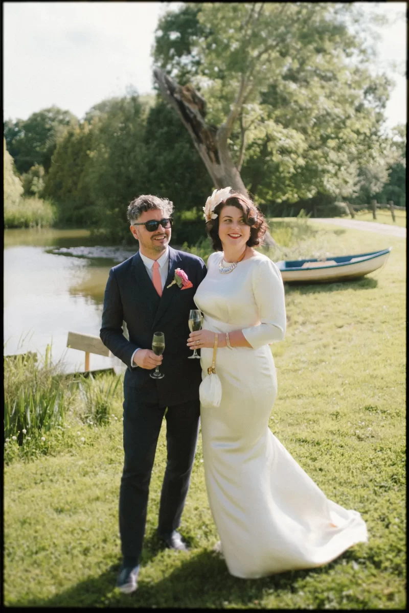 A smiling bride in a white dress and a groom in sunglasses stand together holding drinks on a grassy lawn at their Yoghurt Rooms wedding festival, with a pond, boat, and trees providing the perfect backdrop.