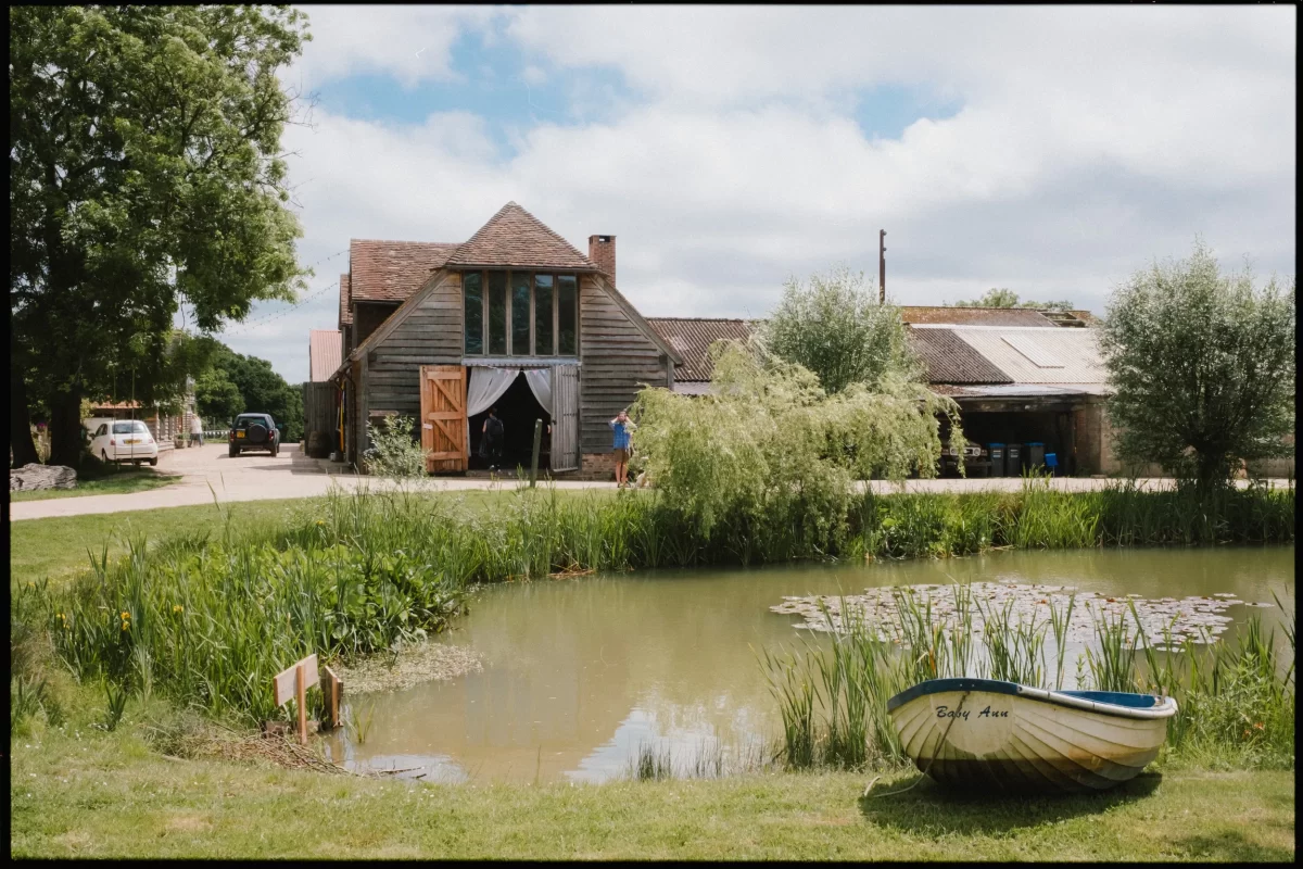 A small pond with lily pads sits in front of rustic wooden barns, perfect for a yoghurt rooms wedding festival. A rowboat labeled Baby Ann rests on the grassy shore, while a person stands near the open barn door amidst trees and greenery.