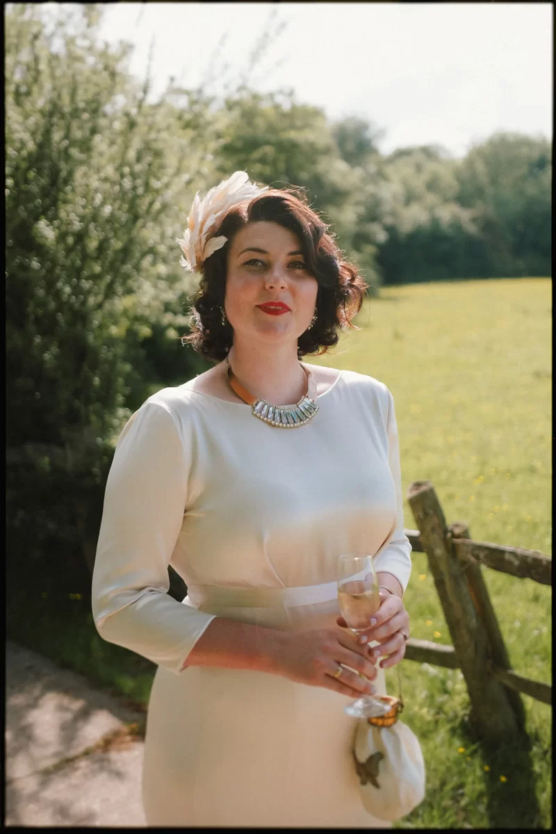 A woman in a white dress and feathered headpiece stands outdoors at a Yoghurt Rooms wedding festival, holding a glass of champagne and a small purse. She wears a statement necklace and red lipstick, with green fields and trees in the background.
