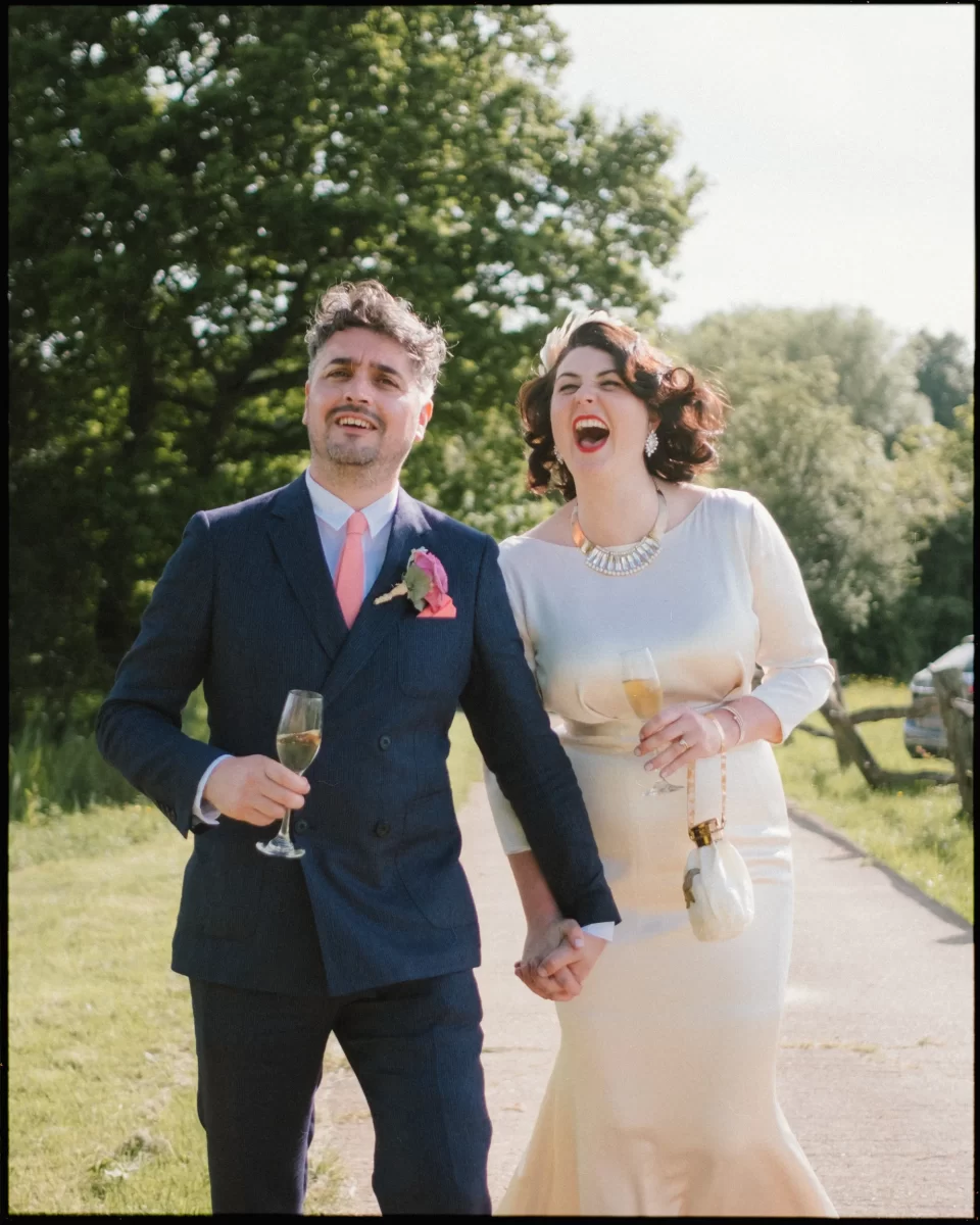 A joyful couple dressed in formal attire laugh and hold hands, strolling outdoors at a Yoghurt Rooms wedding festival, each with a glass of champagne. Green trees and grass surround them on this sunny day.